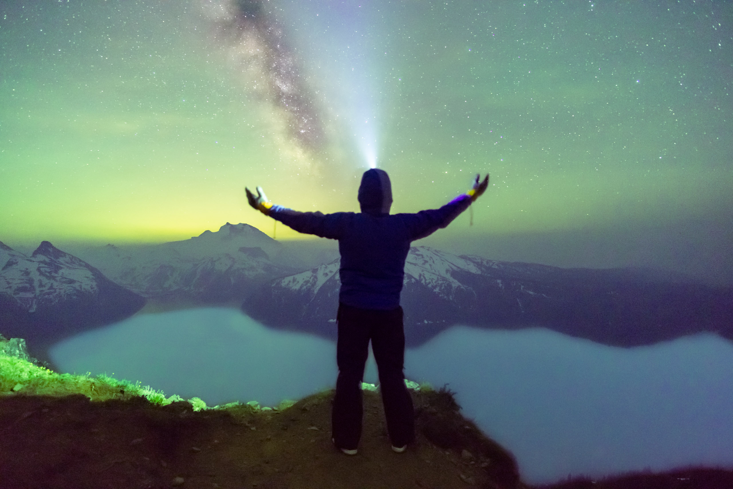 Contact. A man stands on a mountain ridge looking out over a lake a night. The sky is lit by the northern lights and the milkyway.